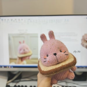 Puede incluir: Un juguete de conejo de punto rosa que lleva un disfraz de pan de punto marrón y beige. El conejo tiene ojos negros y una nariz negra. El juguete está siendo sostenido en una mano.