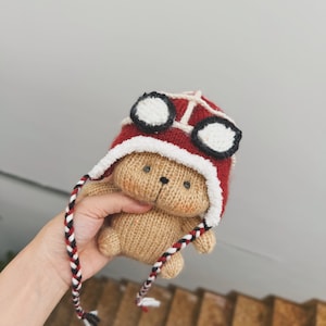 May include: A brown knitted teddy bear wearing a red knitted hat with white and black goggles. The hat has a white fur trim and a long red, white, and black braided cord.