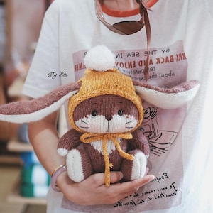 May include: A handmade knitted bunny doll in brown, white, and yellow. The bunny wears a yellow hat with a white pom-pom and has long ears. The doll is held in someone's hands. The text on the shirt reads "KICK BACK AND ENJOY".