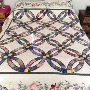 May include: A white quilt with a double ring pattern in blue, yellow, and green. The quilt is decorated with embroidered flowers and birds.