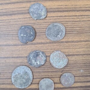 Puede incluir: Una colecci&oacute;n de ocho monedas antiguas, algunas con una p&aacute;tina verdosa, dispuestas en un patr&oacute;n triangular sobre una superficie de madera.