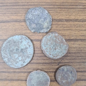 Puede incluir: Cinco monedas antiguas con una p&aacute;tina verdosa sobre una superficie de madera. Las monedas son redondas y tienen un dise&ntilde;o en relieve.