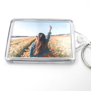 May include: Clear rectangular keyring featuring a photograph of a person in a field, wearing sunglasses and a denim jacket, with their arm raised in a peace sign. The keyring has a silver metal ring.