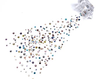 Pode incluir: Um recipiente transparente com uma variedade de strass multicoloridos e iridescentes que se espalham. Os strass são pequenos e redondos, e estão espalhados sobre um fundo branco.