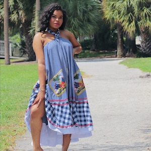 May include: A blue halter dress with a checkered pattern at the hem and decorative patches. The dress has a high slit and is worn with sandals. The background features palm trees and a pathway.