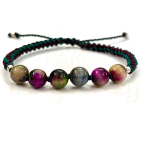 May include: A handmade bracelet featuring a series of colorful, round beads in shades of pink, green, gray, and tan. The beads are strung on a dark green and maroon braided cord, with silver-colored end caps. The bracelet is displayed against a white background.