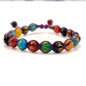 May include: A beaded bracelet featuring colorful round beads with gold lettering, strung on a purple cord. The beads are in various colors, including red, blue, yellow, green, and orange. The bracelet is displayed on a white surface.