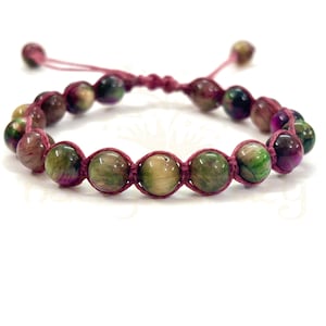 May include: A beaded bracelet featuring a series of round, multi-colored beads in shades of green, purple, and brown. The beads are strung on a pink cord, with adjustable sliding knots for a custom fit. The bracelet is designed for everyday wear.