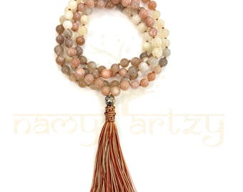 May include: A beaded necklace with a tassel. The necklace is made of small, round, light-colored beads in shades of pink, beige, and gray. The tassel is brown and red. The necklace is displayed on a white background.