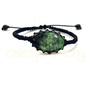 Pode incluir: Um bracelete de cordão preto com uma gema oval verde. A gema está engastada em um envoltório de cordão preto.