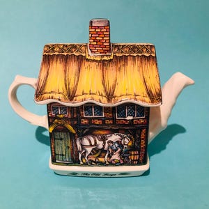 May include: A ceramic teapot shaped like a cottage with a thatched roof and a chimney. The teapot is decorated with a scene of a horse and a person in front of a building. The text "The Old Forge" is printed on the base of the teapot.