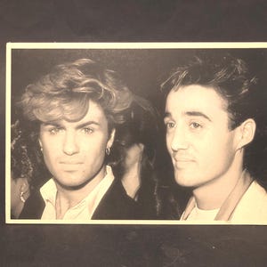 May include: Monochrome photograph of two men. The man on the left has wavy hair and is wearing a dark blazer over a white shirt. The man on the right has styled hair and is wearing a light-coloured shirt. Both men are looking towards the camera.