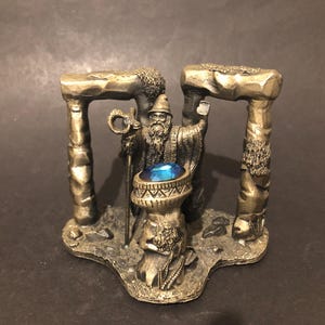 May include: A detailed figurine of a wizard with a long beard and staff, standing between two stone archways. The wizard holds up his hand, and a blue gem sits atop a pedestal. The figurine is made of a metallic material.
