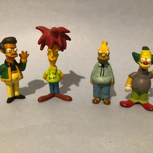 May include: Four Simpsons character figurines. Apu, Sideshow Bob, Homer, and Krusty the Clown are depicted. Sideshow Bob's shirt displays "I H8 BART". The figures are brightly coloured and stand on a light grey surface.