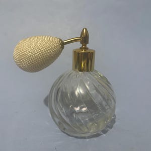 May include: A clear glass perfume bottle with a spherical, spiralled ribbed design. The bottle features a gold-toned metal collar and a matching gold-toned nozzle connected to an oval-shaped, cream-coloured fabric-covered atomiser bulb.