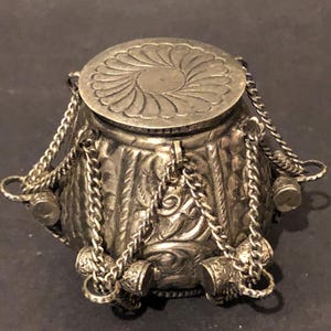 May include: A small, silver-coloured metal container with a lid. The lid features a floral design. Chains hang from the container, and small decorative cylinders are attached to the bottom. The container has an ornate, textured surface.