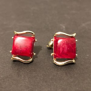 May include: A pair of gold-coloured cufflinks featuring square, deep red centres. The cufflinks have a curved design on the top and bottom, with small decorative accents on each corner. The background is a dark, neutral colour.