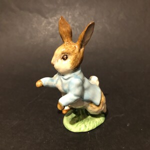 May include: A porcelain figurine of Peter Rabbit from Beatrix Potter's children's book. The figurine depicts Peter Rabbit wearing a blue jacket and standing on a green base.
