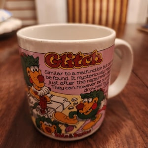 May include: White ceramic mug with a pink band featuring the word "Glitch" and cartoon characters. The mug has a white handle and a humorous description printed on it.