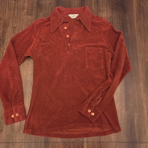 May include: A long-sleeve shirt in a deep rust-orange color, crafted from a soft velour fabric. It features a pointed collar, a three-button placket, a single chest pocket, and buttoned cuffs. The shirt has a straight hem and a visible brand label.