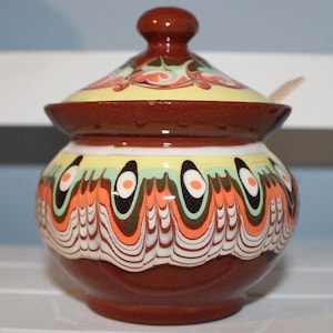 Puede incluir: Un azucarero de cerámica marrón con tapa y una cuchara de madera. El azucarero está decorado con un patrón floral colorido y un diseño geométrico blanco y negro.