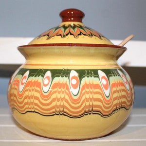 Op de afbeelding: Een gele keramische suikerpot met een deksel en een kleine houten lepel. De kom is versierd met oranje, groene en witte patronen. Het deksel is bruin met een bloemmotief.