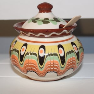 Puede incluir: Un azucarero de cerámica con tapa y una cuchara de madera. El azucarero está decorado con un patrón geométrico colorido en tonos de amarillo, naranja, verde y negro. La tapa tiene una parte superior marrón y una base blanca con un diseño de hojas verdes.