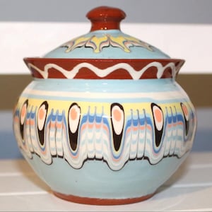 Può includere: Un vaso in ceramica con coperchio, di colore azzurro chiaro, decorato con motivi colorati in giallo, bianco e nero. Il coperchio ha un pomello marrone. L'insieme presenta un aspetto decorativo e artistico.