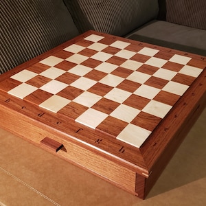 May include: A wooden chessboard with a drawer for storing pieces. The board is made of light and dark wood, and the squares are numbered 1-8 and lettered a-h.