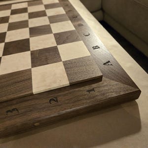 May include: A wooden chessboard featuring a classic alternating pattern of dark brown and light cream-coloured squares. The board is framed by a dark wood border engraved with the letters A through E and the numbers 1 through 3 along the edges.