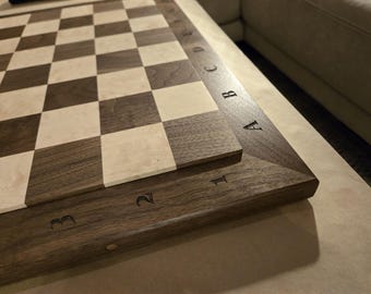 Puede incluir: Un tablero de ajedrez de madera con un patrón clásico de cuadrados alternos en color marrón oscuro y crema claro. El tablero está enmarcado por un borde de madera oscura grabado con las letras A a E y los números 1 a 3 en los bordes.