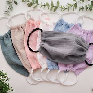 May include: A set of eight face masks made of soft, textured gauze fabric. The masks are in various colours, including light blue, beige, pink, light blue, lavender, and grey. Each mask has a white elastic band and a black elastic band for securing the mask to the face.