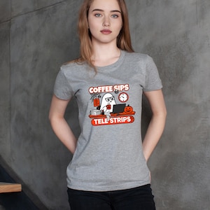 May include: A heather gray short-sleeve t-shirt featuring a graphic of a ghost wearing glasses at a desk with a computer, a pumpkin, a clock, and a spiderweb. The text reads COFFEE SIPS TELE STRIPS in orange and white lettering on the front.