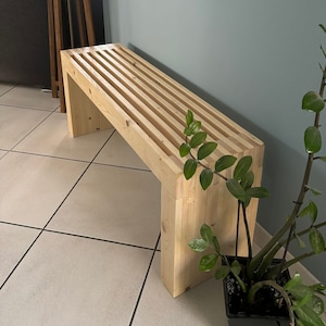 Può includere: Una panca in legno chiaro con un design a doghe, posizionata in un corridoio con una pianta in un vaso nero e una lampada da terra.
