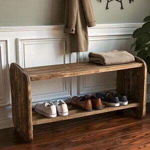 May include: A rustic wooden shoe bench with two shelves. The top shelf holds a folded blanket. Several pairs of shoes are on the lower shelf. A coat hangs above the bench. A potted plant is to the right.