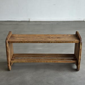 May include: A rustic, two-tiered wooden shoe rack. The rack is constructed from stained wood, showcasing the natural grain. It features a top shelf and a lower shelf with two horizontal slats for shoe storage. The design is simple and functional.