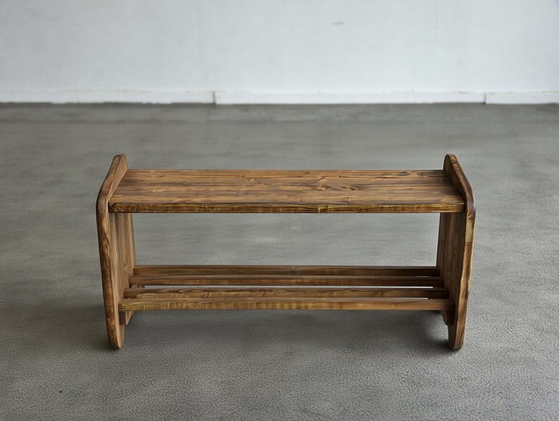 May include: A rustic, two-tiered wooden shoe rack. The rack is constructed from stained wood, showcasing the natural grain. It features a top shelf and a lower shelf with two horizontal slats for shoe storage. The design is simple and functional.
