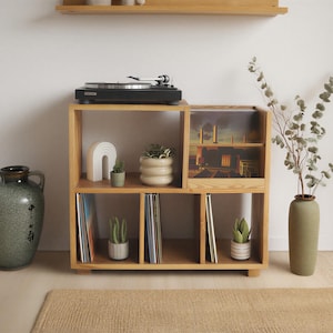 Może przedstawiać: Jasna drewniana półka w kształcie kostki z gramofonem na górze, zielonym wazonem i dużym ceramicznym dzbanem. Półka mieści płyty winylowe, rośliny ozdobne i oprawiony obraz.