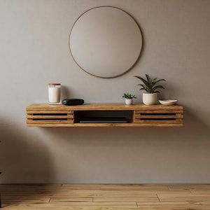 Può includere: Una mensola in legno sospesa con un vano integrato e due cassetti a fessura. Uno specchio rotondo è appeso sopra la mensola. La mensola è decorata con una candela, un piccolo oggetto nero, due piante in vaso e una piccola ciotola. Il muro è dipinto di un colore neutro.