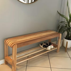 Un banc à chaussures en bois à lattes disposé le long d'un mur gris dans une entrée, avec une paire de chaussures de sport sur l'étagère inférieure.