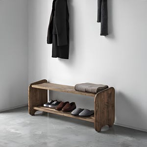 May include: Wooden entryway bench with two shelves. The top shelf holds a folded blanket. The bottom shelf displays several pairs of shoes. A black coat hangs on the wall above the bench. The bench is made of a light brown wood.