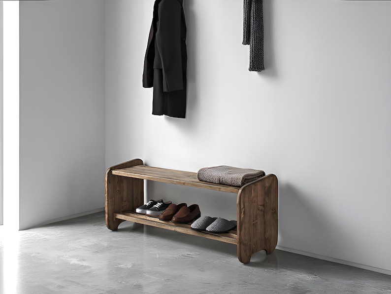 May include: Wooden entryway bench with two shelves. The top shelf holds a folded blanket. The bottom shelf displays several pairs of shoes. A black coat hangs on the wall above the bench. The bench is made of a light brown wood.