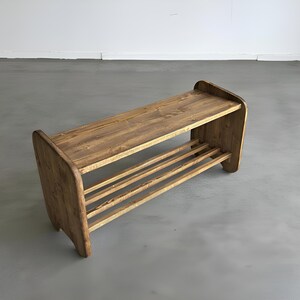 May include: A wooden shoe rack with a natural finish. The rack has a top shelf and a lower shelf with three horizontal slats for shoe storage. The sides of the rack are curved, and the wood grain is visible.