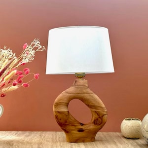 Op de afbeelding: Een houten tafellamp met een witte lampenkap. De lampvoet is gesneden met een uniek ontwerp en heeft een gat in het midden. De lamp staat op een houten tafel.