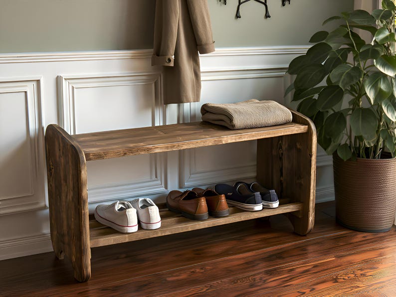 May include: A rustic wooden shoe bench with two shelves. The top shelf holds a folded blanket. Several pairs of shoes are on the lower shelf. A coat hangs above the bench. A potted plant is to the right.
