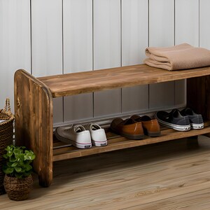 May include: A rustic wooden shoe rack with two shelves, holding several pairs of shoes. A woven basket and a small potted plant sit nearby. A folded beige blanket rests on the top shelf. The rack is made of stained wood.