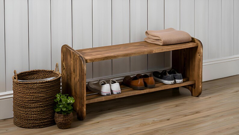 May include: A rustic wooden shoe rack with two shelves, holding several pairs of shoes. A woven basket and a small potted plant sit nearby. A folded beige blanket rests on the top shelf. The rack is made of stained wood.