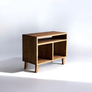 Może przedstawiać: Mała, drewniana szafka do przechowywania z naturalnym wykończeniem. Szafka ma dwie otwarte przegrody i półkę, wspartą na czterech krótkich nogach. Widoczne usłojenie drewna. Na białym tle.