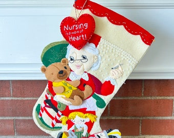 May include: A white felt Christmas stocking with a red heart that says "Nursing is a work of heart!"  The stocking is decorated with a woman holding a teddy bear, a girl with blonde hair, and a nutcracker.