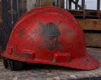 May include: A weathered red hard hat featuring a black graphic of a skull wearing a smaller hard hat, positioned in front of crossed tools including a screwdriver and pliers. The helmet surface shows signs of wear with scuffs and dark marks.